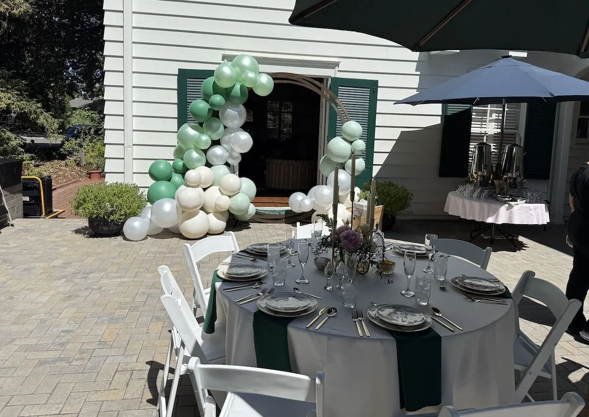 Sage and ivory balloon garland framing a doorway above a table styled with linens and fresh stems — balloon and bloom styling for Campbell catering